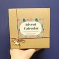 A hand holding a kraft Blue Teapot box tied with red and green twine and a wooden bell. The label reads Advent Calendar, Mixed, 25 teas of 8 g each, 25 paper tea bags, 200 g.