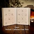 Open white drawer calendar numbered 1 to 24 with small stars, captioned New! Herbal: Caffeine-Free Teas, on a wooden table by a fire.