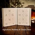 Open white drawer calendar numbered 1 to 24 with small stars, captioned Signature: Holiday & Classic Teas, on a wooden table by a fire.