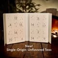Open white drawer calendar numbered 1 to 24 with small stars, captioned New! Single-Origin: Unflavored Teas, on a wooden table by a fire.