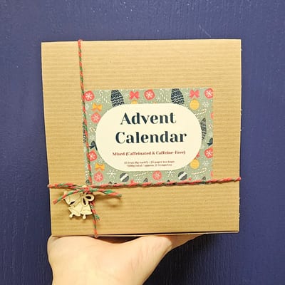 A hand holding a kraft Blue Teapot box tied with red and green twine and a wooden bell. The label reads Advent Calendar, Mixed, 25 teas of 8 g each, 25 paper tea bags, 200 g.