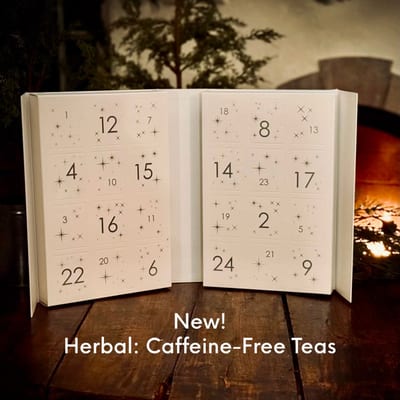 Open white drawer calendar numbered 1 to 24 with small stars, captioned New! Herbal: Caffeine-Free Teas, on a wooden table by a fire.