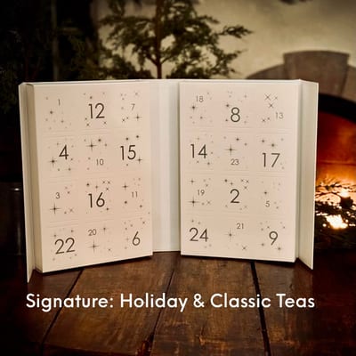 Open white drawer calendar numbered 1 to 24 with small stars, captioned Signature: Holiday & Classic Teas, on a wooden table by a fire.