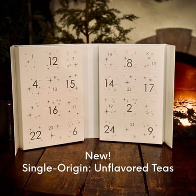 Open white drawer calendar numbered 1 to 24 with small stars, captioned New! Single-Origin: Unflavored Teas, on a wooden table by a fire.