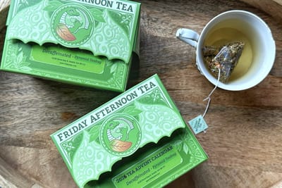Two green Friday Afternoon boxes on a wooden tray, the front one labeled 2026 Tea Advent Calendar, Decaffeinated, Pyramid Teabag, beside a cup with a pyramid bag.