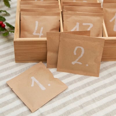 Light wood box of kraft sachets with white painted numbers, sachets marked 1 and 2 lying on a striped cloth beside holly.