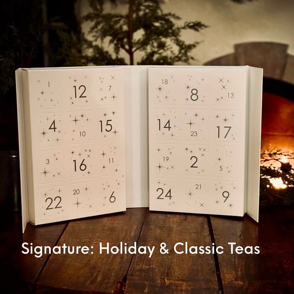 Open white drawer calendar numbered 1 to 24 with small stars, captioned Signature: Holiday & Classic Teas, on a wooden table by a fire.