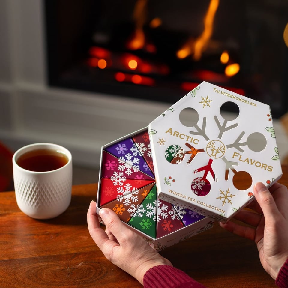Des mains soulèvent le couvercle d'une boîte hexagonale de thés d'hiver Nordqvist près d'une cheminée, une tasse de thé sur la table